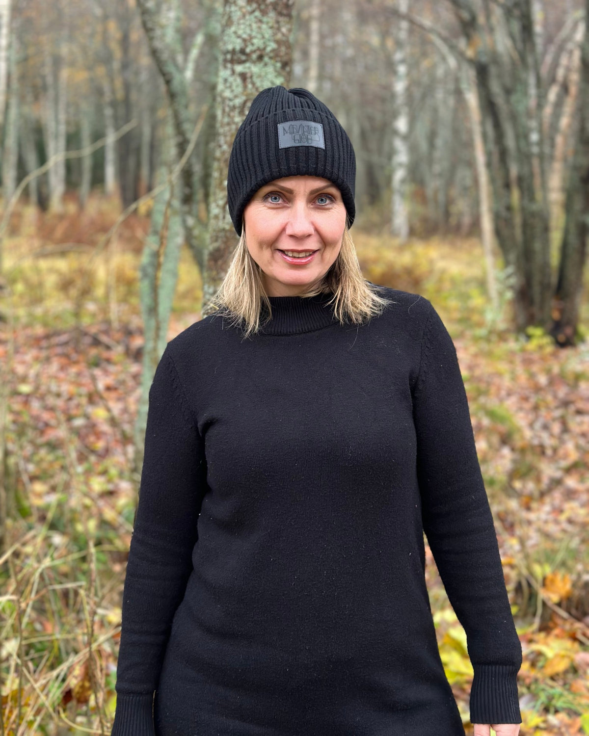 Kvinna i svart tröja står i höstskogen med Midvinterglöd-mössan på huvudet.