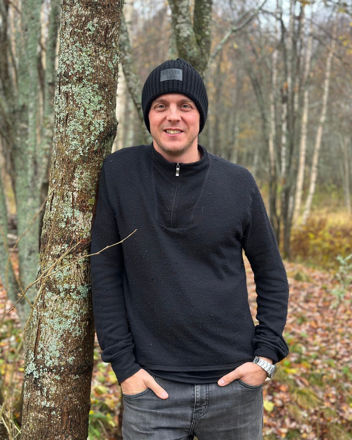 Man lutad mot en björk i höstskogen, bär den svarta Midvinterglöd-mössan.