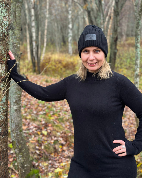 Kvinna med handen mot ett träd i höstskogen, bär Midvinterglöd-mössan.